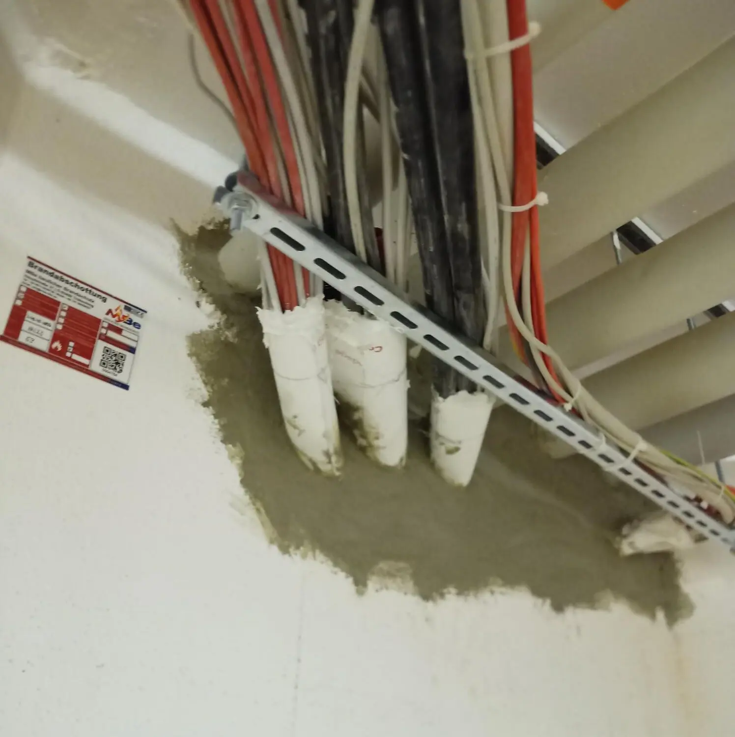 Kabeltrasse, die durch eine Wand geführt und mit Brandschutzmörtel verschlossen ist, daneben das Kennzeichnungsschild der Abschottung.