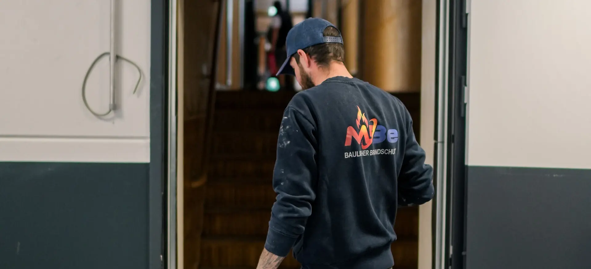 Monteur von MBe Baulicher Brandschutz geht durch eine geöffnete Tür in ein Treppenhaus mit Holztreppe.