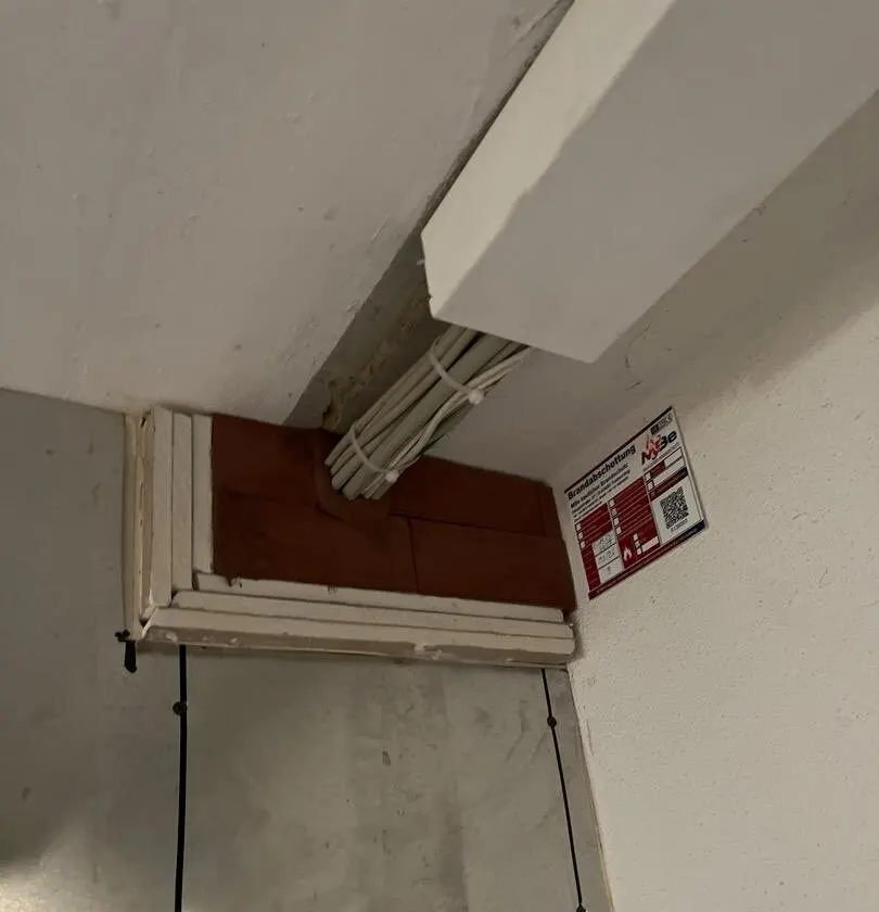 Kabeldurchführung in einer Wand, mit Brandschutzsteinen verschlossen, daneben das Kennzeichnungsschild von MBe Baulicher Brandschutz mit QR-Code.